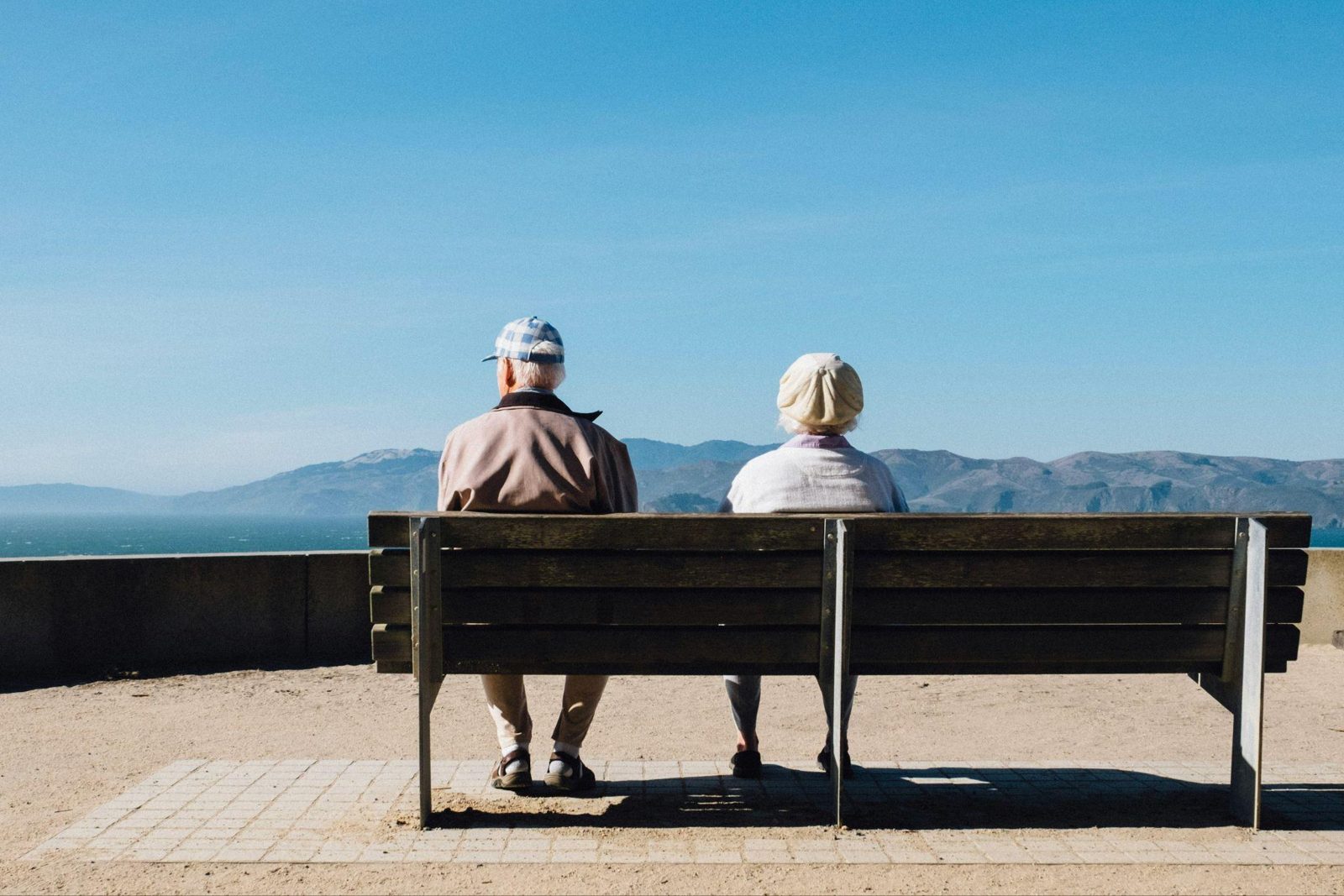 Elderly couple sitting together on a park bench looking out over calm water and distant hills